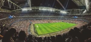 A packed stadium with thousands of spectators watching a soccer match under bright lights. The green field is surrounded by stands filled with fans, and the stadium’s roof is partially visible overhead.