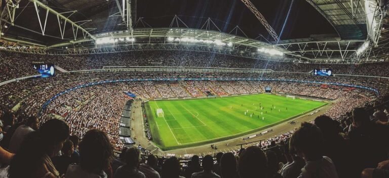 A packed stadium with thousands of spectators watching a soccer match under bright lights. The green field is surrounded by stands filled with fans, and the stadium’s roof is partially visible overhead.