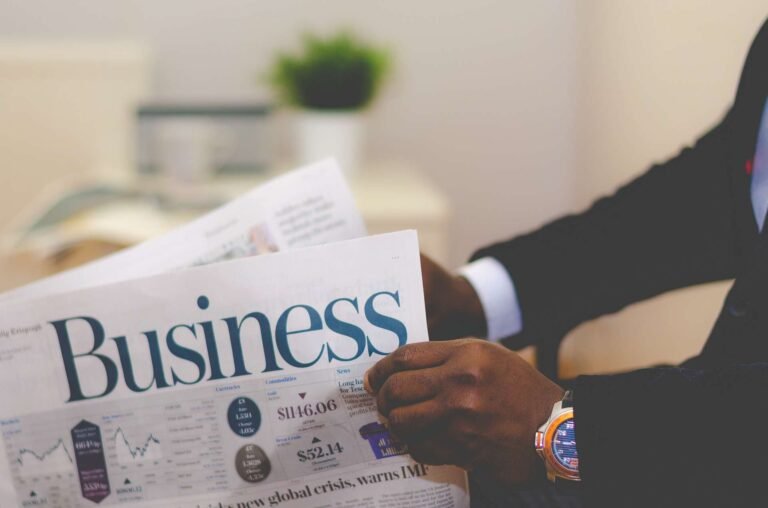A person wearing a suit reads a newspaper with a large Business headline. The individual’s hand and wristwatch are visible, and a blurred office background with a plant can be seen.