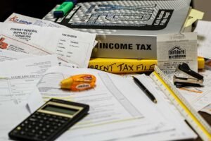 A cluttered desk covered with tax documents, invoices, a calculator, a highlighter, receipts, and an Income Tax binder, representing financial paperwork and tax filing.