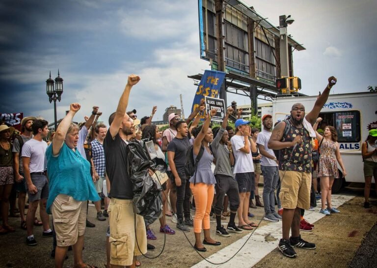 A diverse group of people gathers on a city street, raising fists and holding signs in protest. One person speaks into a microphone, while others look engaged and supportive. A cloudy sky is overhead, and a billboard stands in the background.