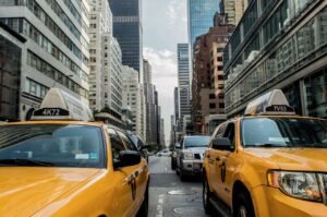 Yellow taxis and cars line a busy street in a city with tall buildings and glass skyscrapers under a partly cloudy sky.
