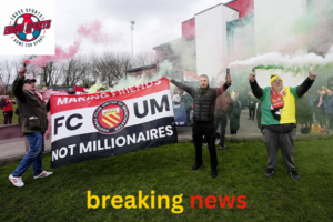 A large crowd of passionate Manchester United supporters gathers outside Broadhurst Park, waving red and yellow anti-Glazer banners.